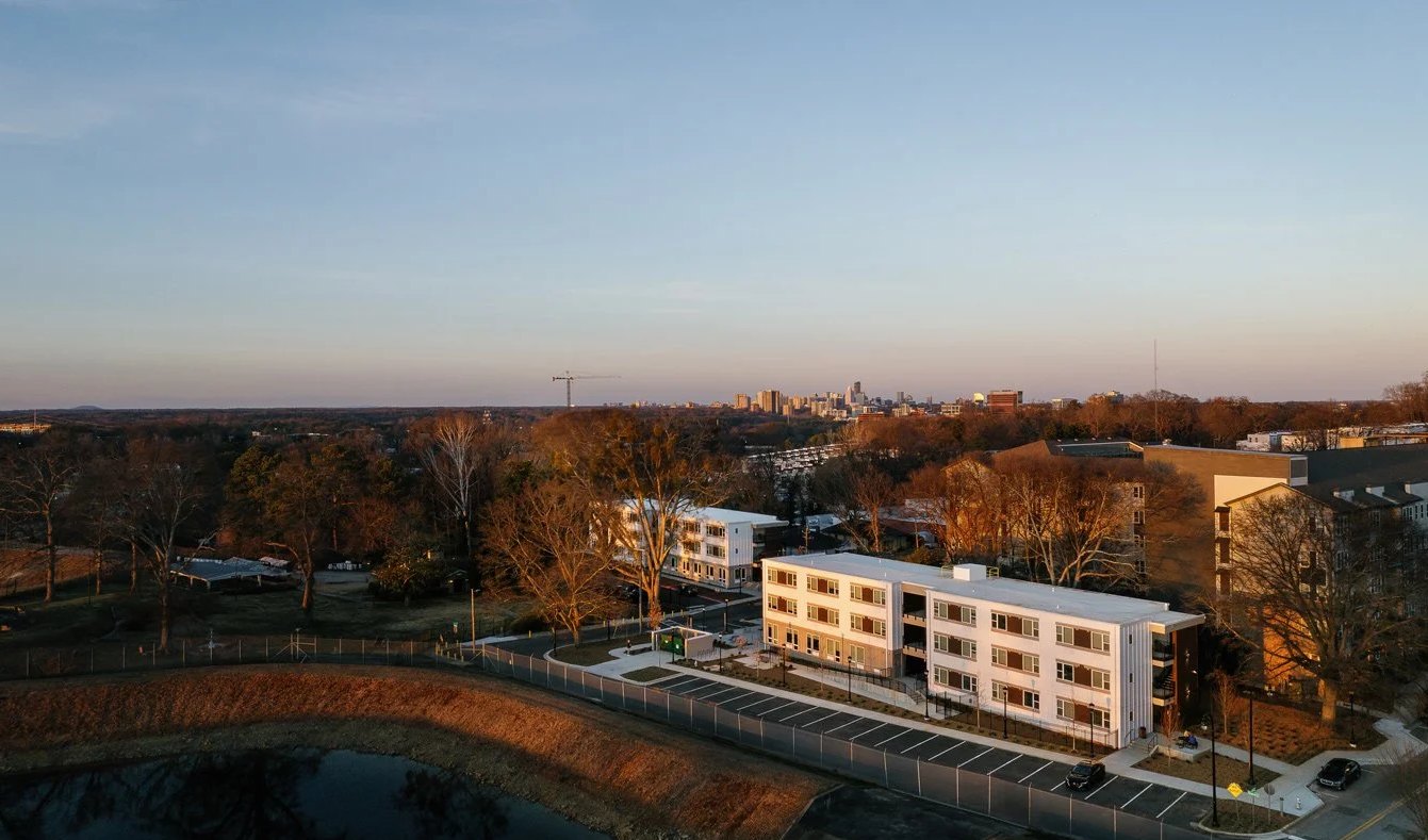 Two three-story apartment buildings at Waterworks Village stand beside a reservoir at sunset, with the Atlanta skyline in the distance.
